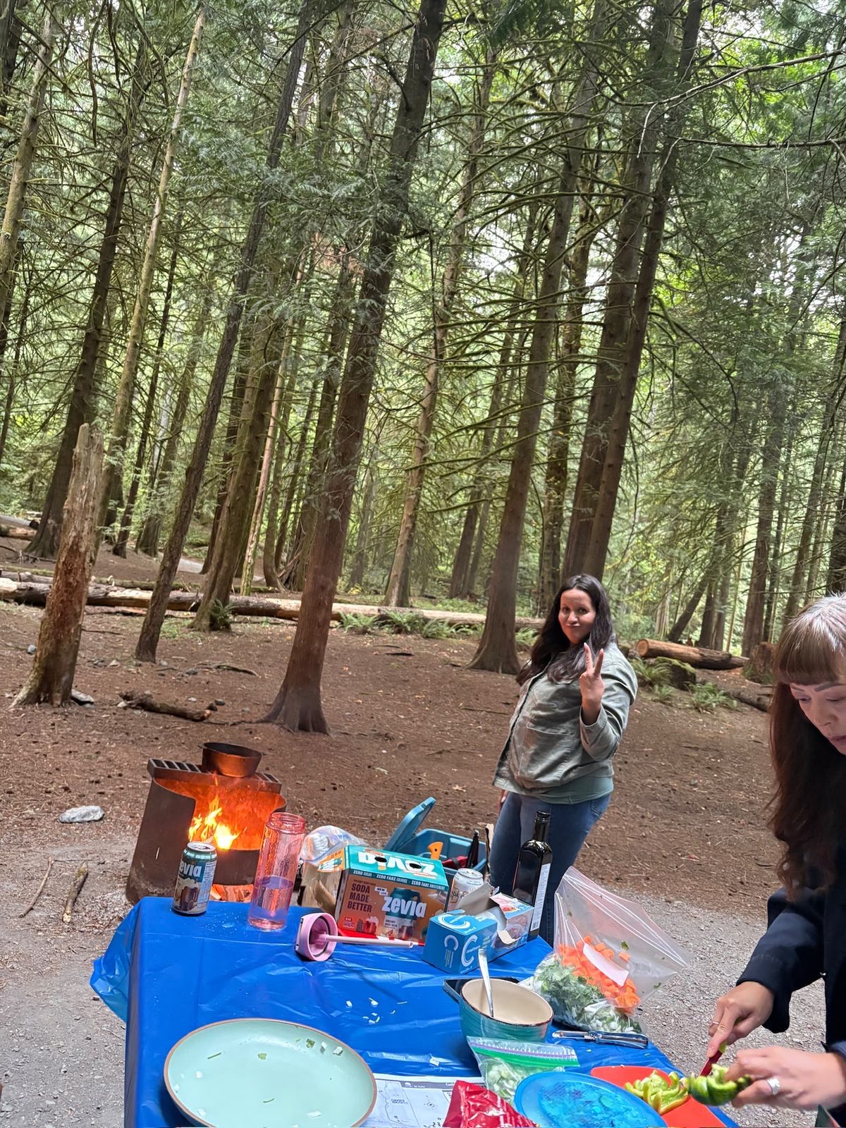 Members setting up the picnic table beside the campfire, one chopping green peppers while another gives a peace sign