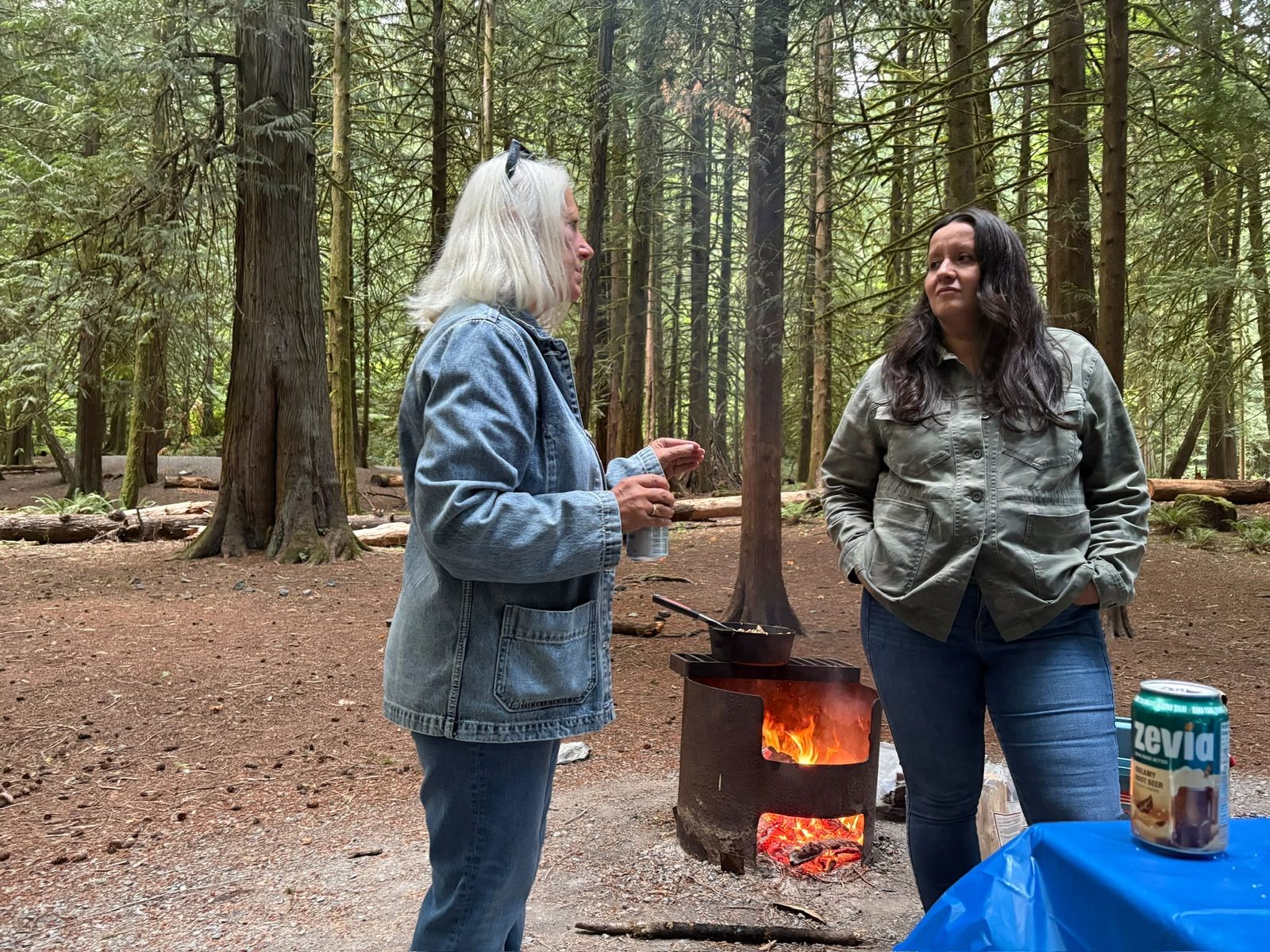 Two members chatting beside the firepit among the tall cedars of Goldstream Provincial Park