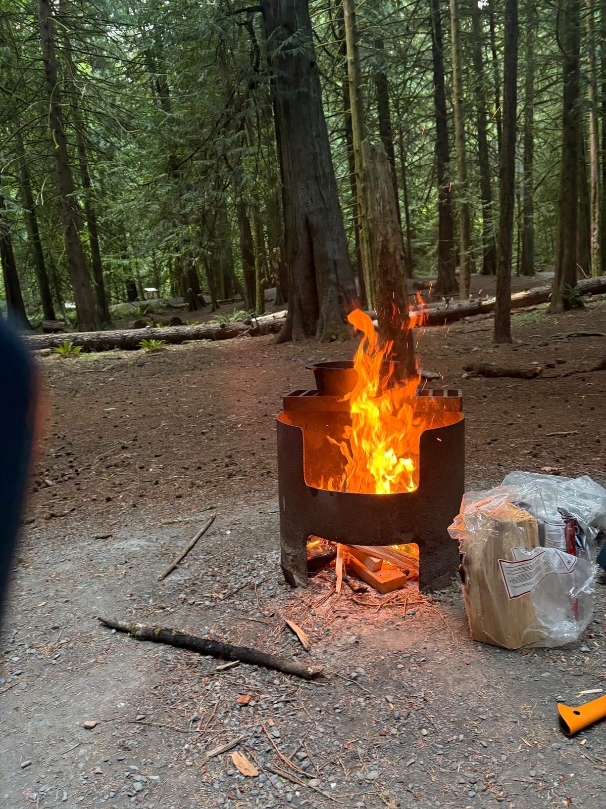 Campfire blazing in the firepit with the Dutch oven on the grate and a bundle of firewood nearby