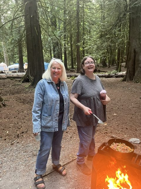 Two smiling members standing by the campfire, one holding a spatula